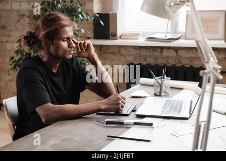 Konzentrierter junger Mixed-Race-Designer mit Pferdeschwanz am Schreibtisch sitzend und mit Zeichentablett beim Erstellen von Maquette auf dem Laptop Stockfoto