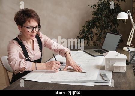 Serious attraktive reife Architekt in Gläsern sitzen am Tisch und mit Dreieck Lineal beim Zeichnen Grundriß im Büro Stockfoto