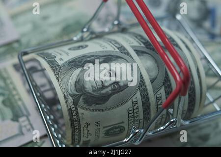 In einen Schlauch gerollte Dollars liegen in einem Einkaufskorb. Der Korb steht auf einem verschwommenen Geldhintergrund. Stockfoto