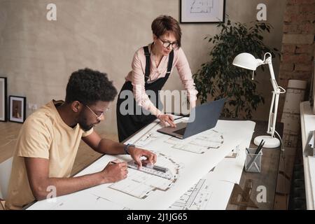 Interracial Gebäude Mitarbeiter arbeiten an Architekturprojekt schwarzer Mann Zeichnung Blaupause, während reife Dame mit Laptop Stockfoto