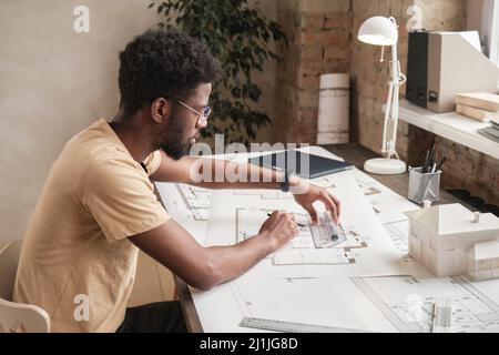 Konzentrierter junger schwarzer Architekt mit Bart, der am Schreibtisch sitzt und ein neues Gebäudedesign kreiert Stockfoto