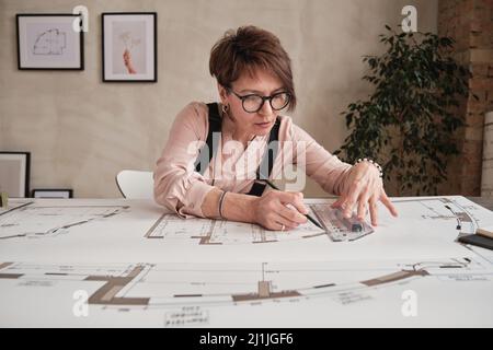 Seriöse reife Frau mit kurzen Haaren sitzt am Tisch im Loft-Büro und mit Lineal, während die Blaupause Zeichnung Stockfoto