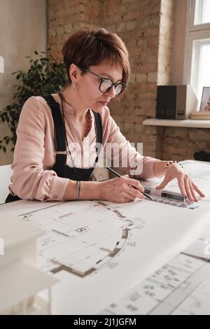 Ernst qualifizierte reife Zeichnerin in Brillen sitzen am Tisch und arbeiten mit Skizze mit Lineal und Bleistift im Büro Stockfoto