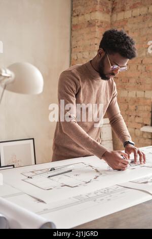 Konzentrierter, schwarz bärtiger Architekt mit Afro-Frisur, der am Schreibtisch steht und im Loft-Büro einen Plan zeichnet Stockfoto