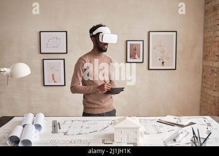 Seriöse junge afroamerikanische Architektin im Pullover, die im modernen Büro mit Bildern an der Wand steht und in einer Virtual-Reality-Brille Maquette zeichnet Stockfoto