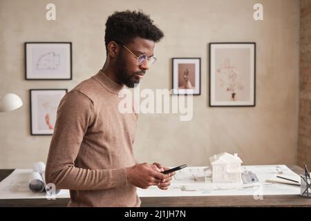 Beschäftigter junger afroamerikanischer Architekt mit Bart, der am Tisch steht, mit Blaupause und SMS an den Kunden Stockfoto