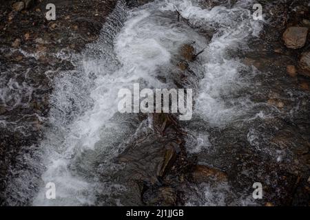 Ein Gebirgsfluss fließt über Steine. Fluss in den Bergen im Winter. Ein Bach aus Wasser entlang eines felsigen Abhangs. Klares Wasser aus einem Gletscher fließt entlang einer Stockfoto