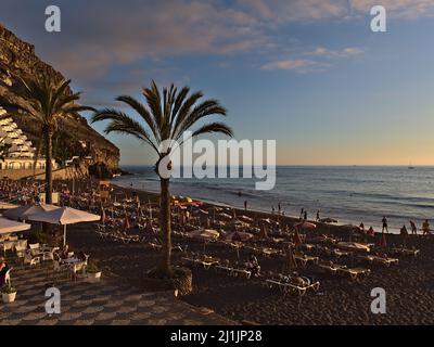 Schöner Blick auf den beliebten Strand von Taurito, Gran Canaria, Spanien, mit Menschen, die die Abendsonne auf Stühlen an der Küste des Atlantischen Ozeans genießen. Stockfoto