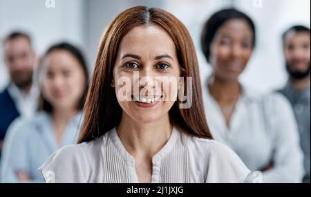 Ich nehme mein Team direkt an die Spitze. Porträt einer Geschäftsfrau, die in einem Büro mit ihren Kollegen im Hintergrund steht. Stockfoto