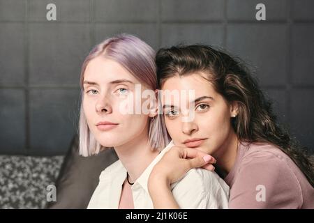 Ruhige junge braunhaarige Frau, die sich auf die Schulter der Freundin lehnt und sie von hinten im Schlafzimmer umarmt Stockfoto