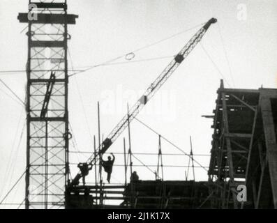 Bauarbeiter halten sich mit einem Kran im Hintergrund auf das Gerüst. Foto von Angelo Rizzuto. 1957. Stockfoto