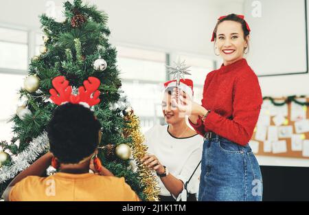 Ein Weihnachtsbaum ist nicht komplett ohne den leuchtenden Stern. Porträt einer attraktiven jungen Geschäftsfrau, die mit ihren Kollegen einen Weihnachtsbaum schmückt Stockfoto
