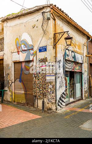 Zwei große Wandgesichter schmücken hier die Rollläden des Eckgebäudes, das mit Kurzwarentafeln an der Yeşil Gazino Sokak in der Altstadt von Lefkoşa (Nikosia), der Türkischen Republik Zypern (TRNK), behangen ist; die Gasse dahinter ist mit rosa und grauem Stein gepflastert. Stockfoto