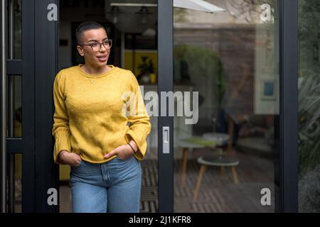 Ruhige Erwachsene Frau, die eine Brille trägt, die Hände in den Taschen hält, während sie in ihrem Hof steht. Stockfoto
