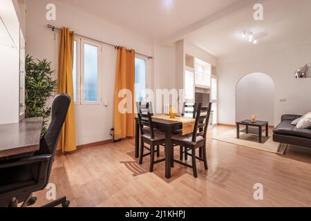 Wohnzimmer mit Esstisch aus dunklem Holz und passenden Stühlen mit gelben Akzenten, die zu den Vorhängen passen, Holzschreibtisch und schwarzem Bürostuhl Stockfoto