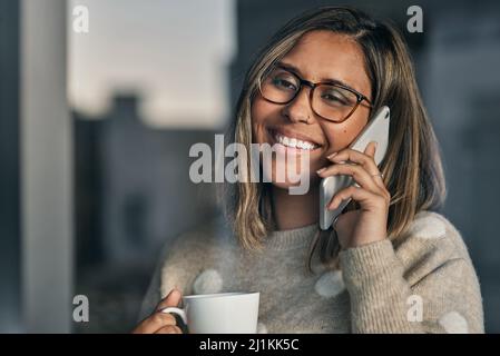 Ich habe auf Ihren Anruf gewartet. Aufnahme einer Frau, die eine Tasse Kaffee in der Hand hält, während sie zu Hause auf ihrem Handy spricht. Stockfoto