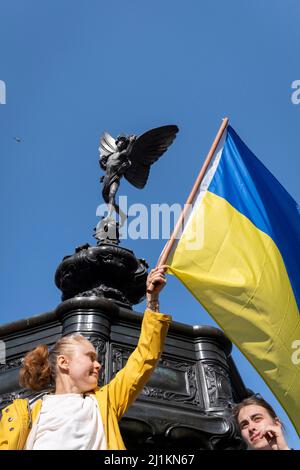 Eine ukrainische Frau hält ihre Nationalflagge unter der Statue des Eros im Piccadilly Circus während des Anti-Kriegs-protestmarsches durch das Zentrum Londons gegen die russische Invasion in der Ukraine am 26.. März 2022 in London, England. Der durchmarsch wurde vom Londoner Bürgermeister Sadiq Khan organisiert. Stockfoto