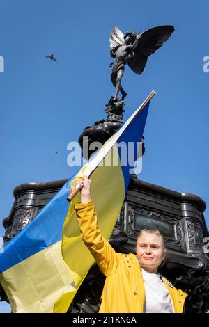 Eine ukrainische Frau hält ihre Nationalflagge unter der Statue des Eros im Piccadilly Circus während des Anti-Kriegs-protestmarsches durch das Zentrum Londons gegen die russische Invasion in der Ukraine am 26.. März 2022 in London, England. Der durchmarsch wurde vom Londoner Bürgermeister Sadiq Khan organisiert. Stockfoto