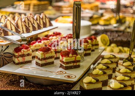 Kleine geschichtete Kuchenstücke mit einem Schokoladenbiskuitboden und einer hellen Sahneschicht, belegt mit frischen Erdbeeren, sind auf einem Tablett am Dessertbuffet aufgereiht, vermutlich im Salamis Bay Conti Hotel in der Nähe von Yeni Boğaziçi, Türkische Republik Zypern (TRNC). Dreieckige Gebäckstücke warten im Hintergrund, während rechts weitere Kuchenquadrate jeweils mit einem Klecks heller und dunkler Schokolade belegt sind. Stockfoto