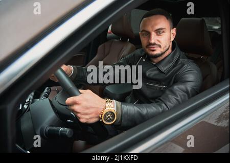Porträt eines hübschen jungen Mannes, der ein modernes Auto fährt. Typ in legerer Kleidung und goldener Uhr. Stockfoto
