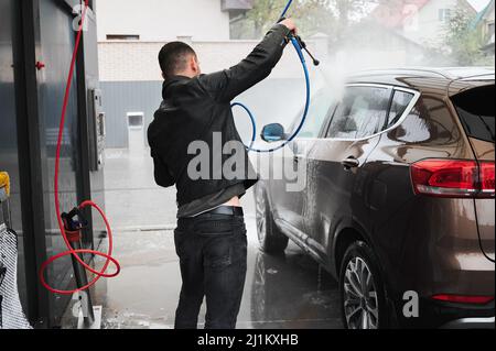 Rückansicht des Besitzers eines Luxuswagens, der den Transport mit fließendem Druckwasser aus der Pistole wäscht. Kfz-Reinigung mit professioneller Ausrüstung bei Selbstbedienungswaschung. Stockfoto