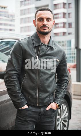 Porträt eines hübschen jungen Mannes, der in der Nähe seines modernen Autos im Freien posiert. Fahrer in legerer Kleidung und goldener Uhr. Stockfoto