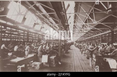 Royal Small Arms Factory, Enfield, Hauptmaschinenfabrik, erster Weltkrieg: Munitionsarbeiterinnen, die auf Arbeitsbänken mit Munitionskisten neben ihnen sitzen. Fotopostkarte, verkauft von Wakefield's, Ealing, London. Stockfoto