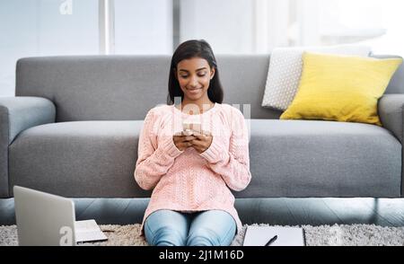 Ich überprüfe nur meine Texte, bevor ich wieder an die Arbeit gehe. Ausgeschnittene Aufnahme einer jungen schönen Frau, die ein Mobiltelefon benutzt, während sie im Wohnzimmer auf dem Boden sitzt Stockfoto