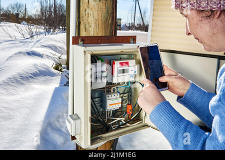 Im Winter nimmt die Frau Messwerte von einem elektrischen Messgerät in einem elektrischen Außenpanel auf und fotografiert das Display des Geräts. Stockfoto