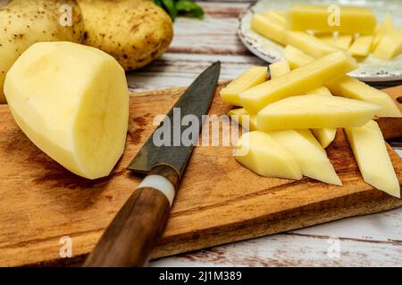 Holzschneidebrett mit einer Kartoffel, die in eine Stabform geschnitten wurde, um Pommes frites zuzubereiten. Eine halbe geschälte Kartoffel und ein Messer auf einem rustikalen Tisch. Hohe Ansicht. Stockfoto