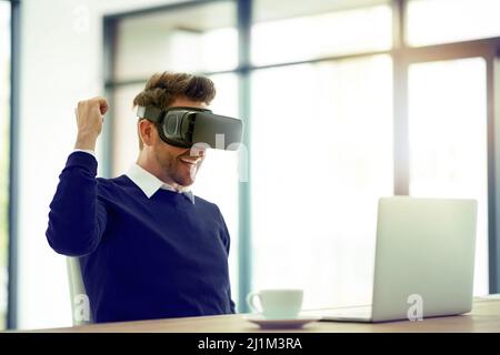 Er hat gerade einen weiteren Sieg erzielt. Aufnahme eines Geschäftsmanns, der ein VR-Headset trägt und während der Arbeit in einem Büro eine Faustpumpe macht. Stockfoto