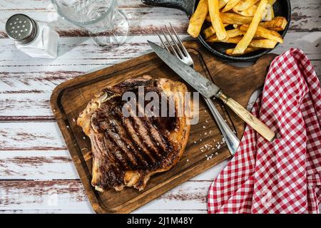 Unglaublich saftiger T-Bone, der auf dem Grill oder Grill auf einem Holzbrett mit Pommes frites und Besteck an der Seite gekocht wird. Draufsicht. Stockfoto