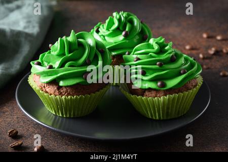 Drei Schokoladen-Cupcakes mit grüner Schlagsahne und Schokoladenstreuseln auf dunkelbraunem Hintergrund. St. Patrick's Day. Nahaufnahme. Stockfoto