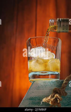 Whiskey aus einer Flasche wird mit Eis in ein Glas gegossen. Holzhintergrund. Alkoholisches Getränk. Moskau Russland 27. März 2022. Stockfoto