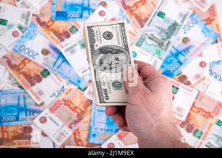 Ein Mann hält vor dem Hintergrund von Rubel verschiedener Konfessionen in Nahaufnahme ein Päckchen mit 100-Dollar-Scheinen aus Stockfoto
