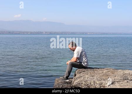 Ein charmanter Mann, etwa 40 Jahre alt, sitzt auf Felsen und starrt auf den See. Stockfoto