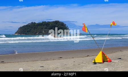 Gekreuzte Flaggen des Surfbrands, die bedeuten, dass man am Strand nicht schwimmen darf. Fotografiert am Mount Maunganui, Neuseeland. Vor der Küste liegt die Insel Motuotau Stockfoto