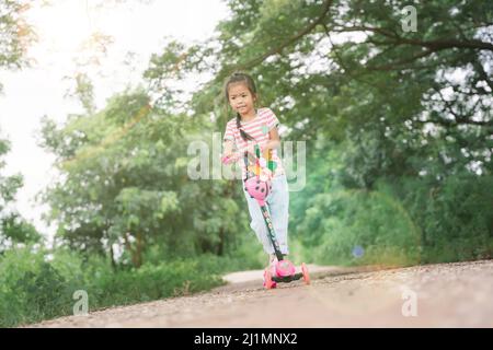 Kinder in Asien lernen an einem Sommertag in einem Park Motorroller zu fahren. Vorschulmädchen, die eine Rolle reitet. Kinder spielen draußen mit Motorroller. Aktive Freizeit und Stockfoto