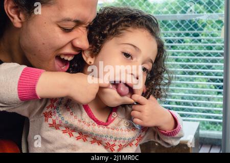 Vater und Tochter lächeln und haben Spaß zusammen in der Familie drinnen, kleines Mädchen ragte aus ihrer Zunge öffnen ihren Mund mit ihren Fingern spielen Stockfoto