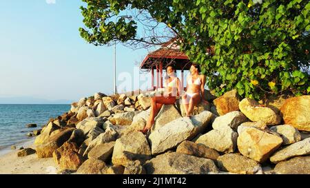 Zwei kaukasische hübsche Frauen im Bikini, die am felsigen Strand in Koh Samui, Thailand, sitzen Stockfoto