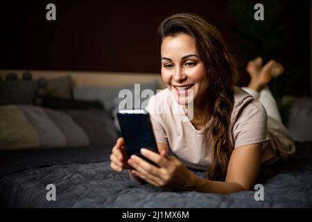 Glückliche Hausfrau im Schlafanzug liegend bequemes Bett chatten Smartphone online Dating-Anwendung Stockfoto