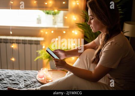 Nachdenkliche moderne Hausfrau im Schlafanzug SMS-Nachricht Smartphone auf dem Bett sitzen Stockfoto