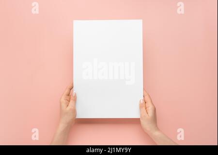 Weibliche Hände halten leeres Papier isoliert auf rosa Hintergrund in vertikaler Position. Frau Hände mit Mockup leeres Papier Blatt auf rosa Hintergrund. Hände Stockfoto