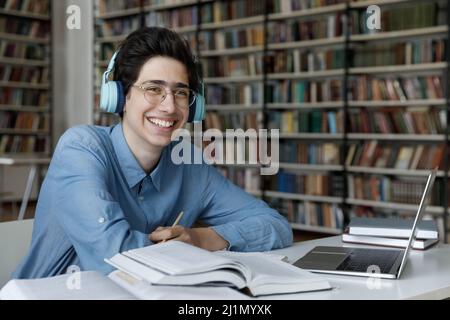 Glücklicher Student Kerl in Kopfhörer sitzen am Tisch in der Bibliothek Stockfoto