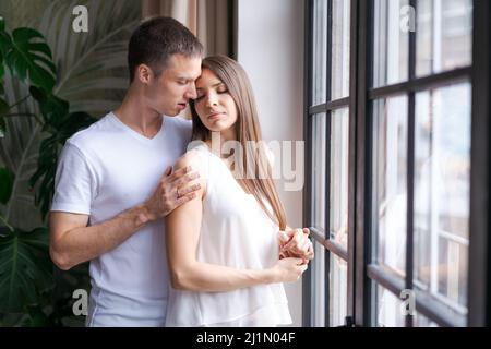 Sehen Sie glückliches tausendjähriges kaukasisches Paar Blick aus dem Fenster Entfernung träumen oder denken. Lächelnde junge Männer und Frauen umarmen Umarmungen feiern den Umzug in Ihr eigenes Zuhause oder Zuhause. Stockfoto