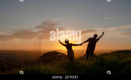 Das Silhouette-Paar steht auf dem Hügel mit offenen Armen und begrüßt die Sonne. One Tree Hill, Auckland. Stockfoto