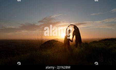 Silhouette eines Paares, das auf einem Hügel steht und die Sonne umarmt. One Tree Hill, Auckland. Stockfoto