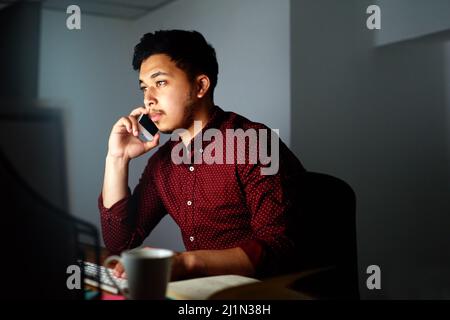 In Kontakt bleiben 247. Aufnahme eines jungen männlichen Designers, der spät in der Nacht am Telefon sprach und an seinem Computer arbeitete. Stockfoto