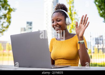 Lächelnde, attraktive junge afrikanerin, die in einem öffentlichen Stadtpark einen Videoanruf abruft, schnurlose Kopfhörertechnologie nutzt, Familie und Freunde begrüßt, Stockfoto