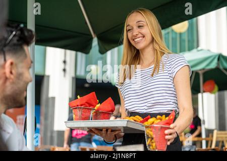 Die junge blonde Kellnerin serviert Pommes Frites und Bier an den Tischen, in der Gastronomie und im Restaurant Stockfoto